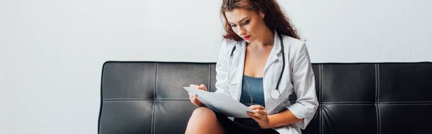 panoramic shot of sexy nurse looking at document and sitting on sofa - Foto, afbeelding