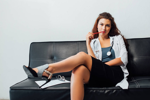 sexy nurse sitting on sofa and holding pencil near lips  - Fotó, kép