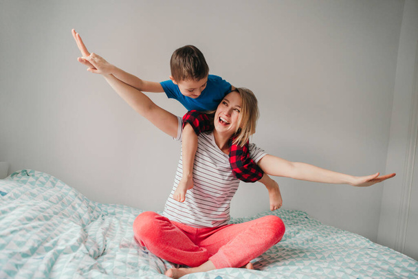 Smiling Caucasian mother and boy son playing in bedroom at home. Child sitting on mom shoulders and laughing. Family having fun together. Happy childhood candid authentic lifestyle. - Valokuva, kuva