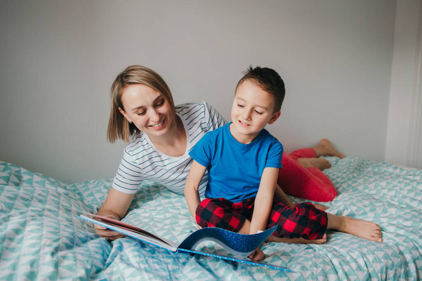 幸せな白人の母親と息子の家族は寝室で本を読んでいます。家で一緒に過ごす母親と少年の子供。親が子供と話してる。本物の人々本物のライフスタイル. - 写真・画像