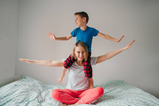 Smiling Caucasian mother and boy son playing in bedroom at home. Child sitting on mom shoulders and laughing. Family having fun together. Happy childhood candid authentic lifestyle. - Photo, Image