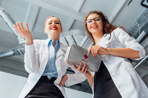 Низкий угол обзора счастливых и сексуальных медсестер в белых халатах с аптечкой
  - Фото, изображение