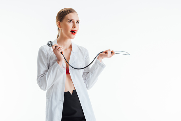 excited sexy nurse in white coat holding stethoscope isolated on white  - Fotografie, Obrázek