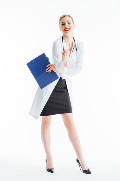 sonriente sexy enfermera sosteniendo portapapeles y pluma mientras está de pie en blanco
  - Foto, Imagen