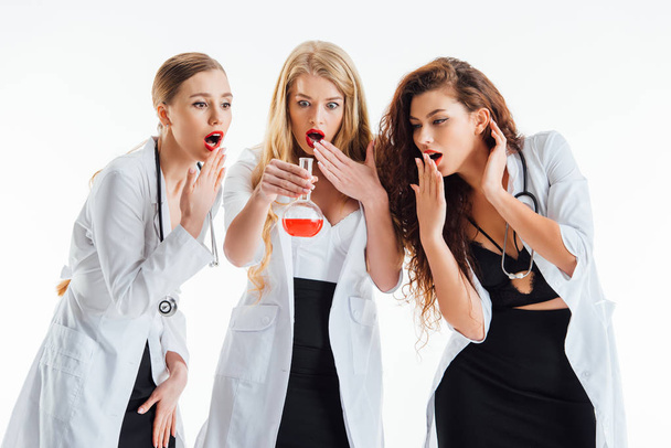 shocked and sexy nurses looking at glass flask with red liquid isolated on white  - Foto, Imagem