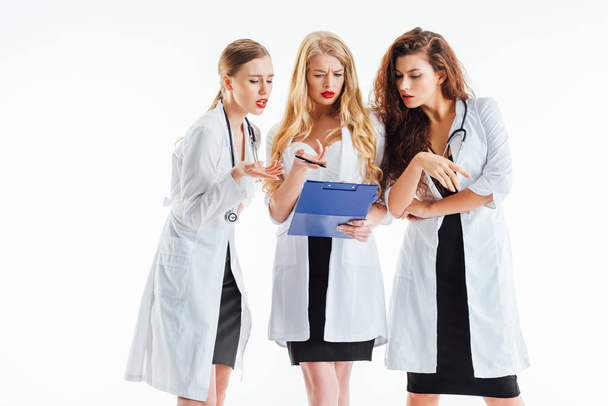 сексуальные медсестры в белых халатах жестикулируют, глядя на блокнот, изолированный на белом
  - Фото, изображение