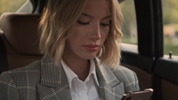 Close up view of Smiling blonde business woman in coat using smartphone while sitting in car - Imágenes, Vídeo
