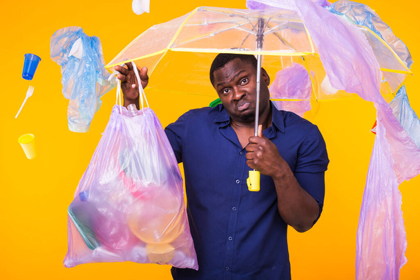 ゴミの問題、プラスチックのリサイクル、汚染と環境の概念-黄色の背景にゴミ袋を運ぶ混乱した男 - 写真・画像