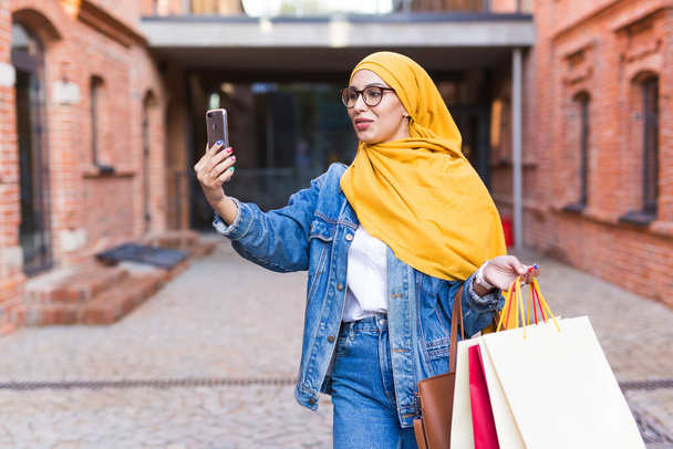 Venta, tecnologías y concepto de compra - Mujer musulmana árabe feliz tomando selfie al aire libre después de ir de compras
 - Foto, Imagen