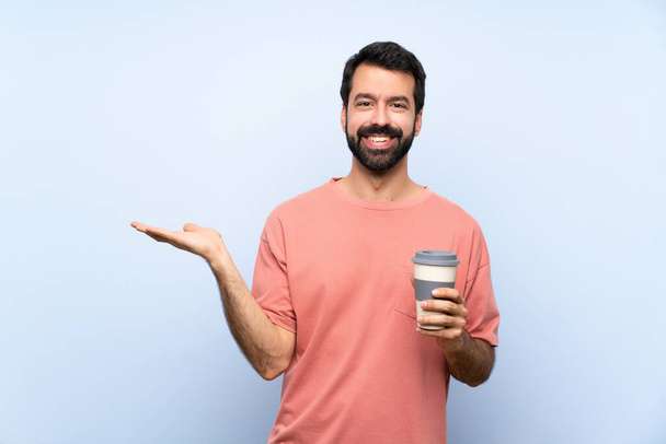Young man with beard holding a take away coffee over isolated blue background holding copyspace imaginary on the palm to insert an ad - Fotoğraf, Görsel