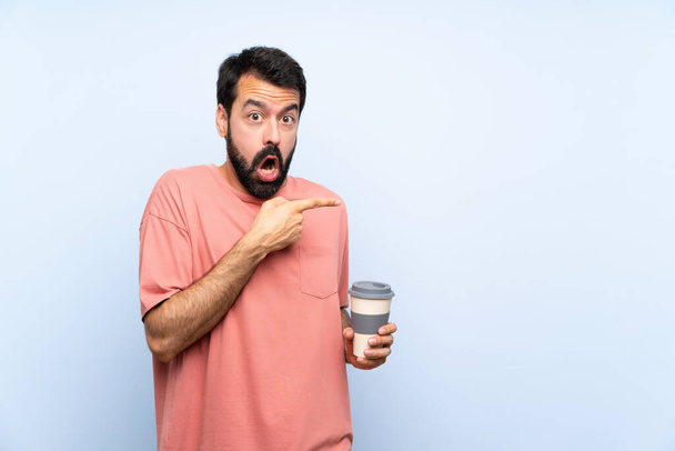 Mladý muž s plnovousem drží odnést kávu přes izolované modré pozadí překvapen a ukazuje stranou - Fotografie, Obrázek