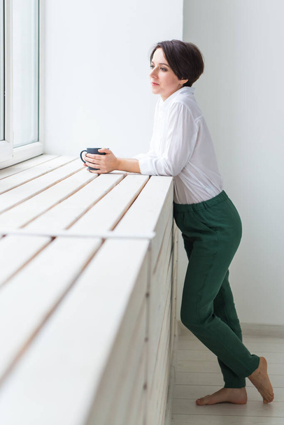 Спокойствие и уют. Красивая молодая женщина с чашкой чая стоя у окна дома
 - Фото, изображение