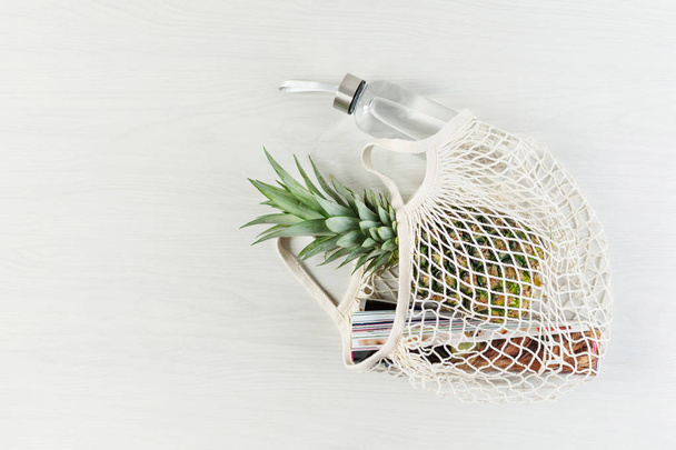 Shopping bag in rete con ananas, rivista lucida e bottiglia d'acqua ecologica su fondo bianco in legno. Stile di vita sostenibile
. - Foto, immagini