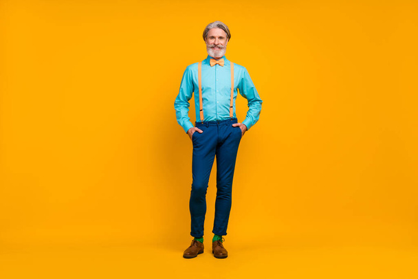 Full length photo of cool trendy grandpa hold hands pockets charismatic person stylish look clothes wear shirt suspenders bow tie pants boots socks isolated yellow color background - Foto, afbeelding