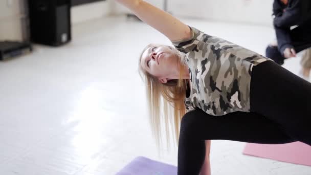 aantrekkelijke blonde vrouw op de voorgrond en groep van jonge sportieve mensen oefenen yoga indoor - Video