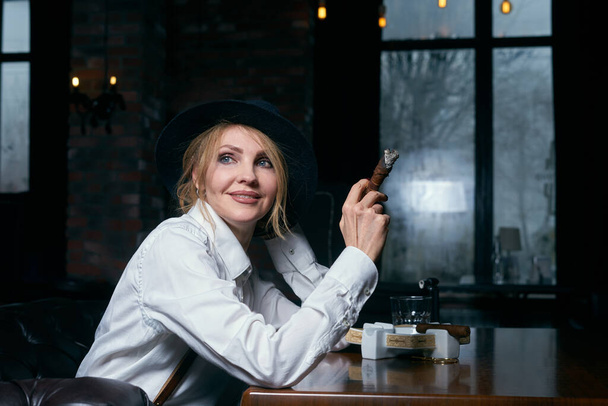 Senior attractive female gangster smoking cigar over dark background in restaurant - Fotoğraf, Görsel
