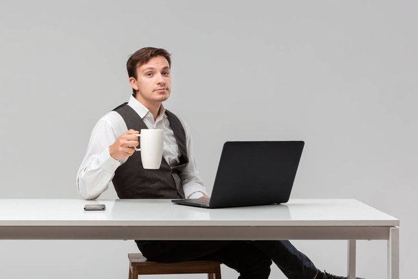 Successful man in a white t-shirt and grey vest drinking coffee sitting at the white table - Fotoğraf, Görsel