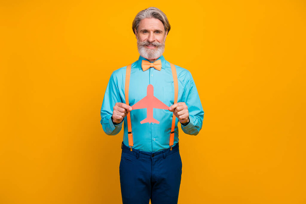 Photo of cool stylish grandpa hold paper air plane recommending choose new airline transportation wear blue shirt suspenders bow tie pants isolated yellow color background - Fotoğraf, Görsel