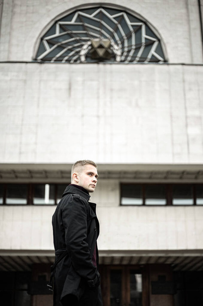 Стильный мужчина в черном плаще и поло на фоне модернистского здания
 - Фото, изображение