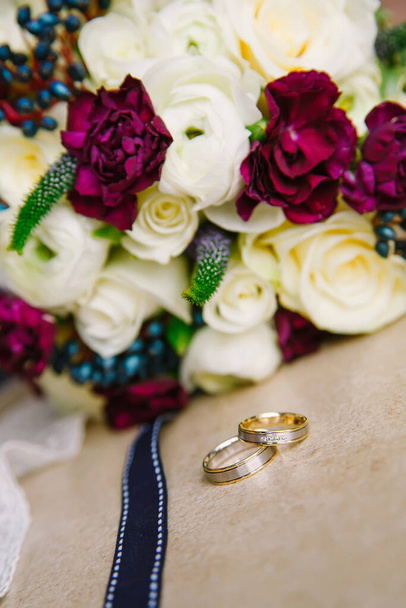 Gold wedding rings lie on the table against the background of a bouquet of flowers - 写真・画像