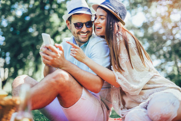 果物でいっぱいのピクニックバスケットがある公園でピクニックブランケットに寝そべっているカップルは、スマートフォンを使用しています - 写真・画像