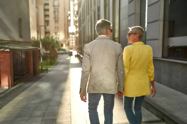 Back view of stylish middle-aged couple holding hands and looking at each other while walking together through the city street - Valokuva, kuva