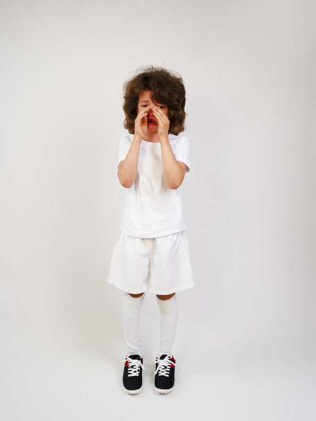 Sports injury. Youth European soccer player in white uniform isolated on gray background. Studio shot. Full body. Boy standing and crying. - Zdjęcie, obraz