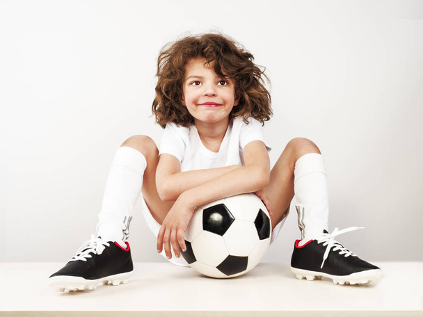 хлопчик з футбольним м'ячем сидить на підлозі на світлому фоні
 - Фото, зображення