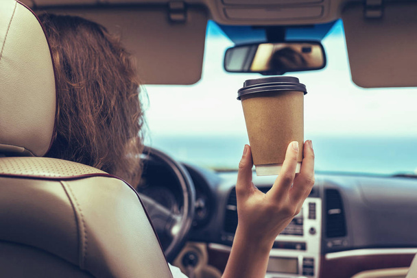 Woman driver drinking coffee paper cup inside car during driving. Girl relaxing in auto trip traveling along ocean tropical beach in background. Traveler concept. Back view - Photo, image