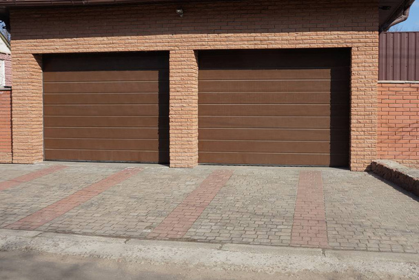 фасад гаража из коричневого кирпича с двумя металлическими воротами на каменном тротуаре на улице в солнечный день
 - Фото, изображение