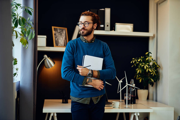 Handsome positive young caucasian hipster standing in his office, holding paperwork and preparing to go on meeting with important clients. - Photo, image