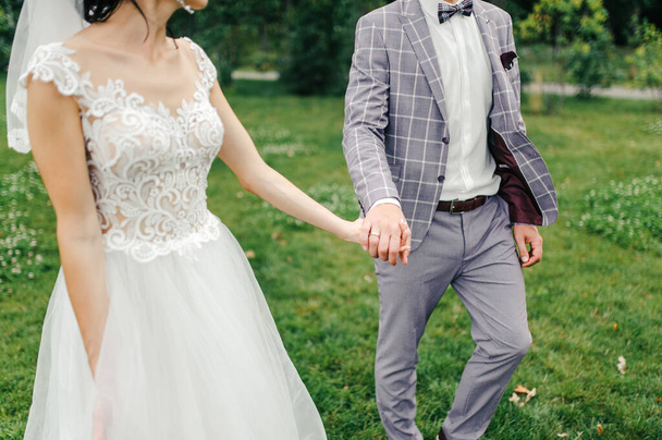 Groom in a grey suit and bride in a wedding dress and veil are walking on a green lawn - Foto, Imagen