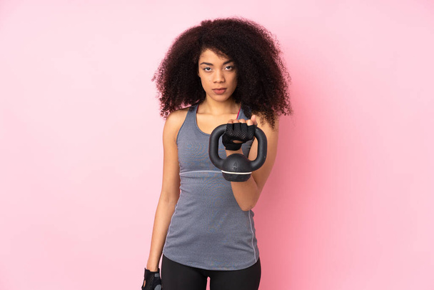 Jovem afro-americana esporte mulher isolada no fundo rosa fazendo levantamento de peso com kettlebell e olhando para a frente
 - Foto, Imagem