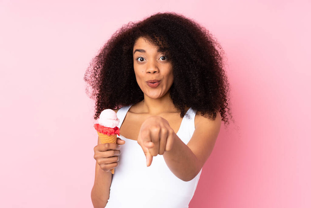 Young african american woman holding a cornet ice cream isolated on pink background surprised and pointing front - Foto, Bild