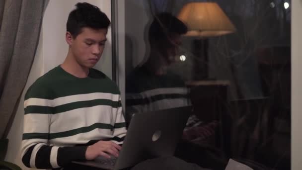 Portrait of young Asian boy typing on laptop and looking out the window in the evening. Handsome college student studying indoors. Ideas, intelligence, online education. - Filmmaterial, Video