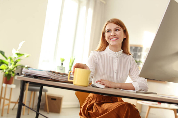 Young woman with cup of drink relaxing at table in office during break - Foto, Bild