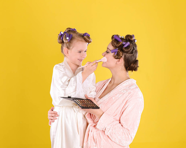 Happy daughter applying powder onto mother's face on yellow background - Foto, Imagen
