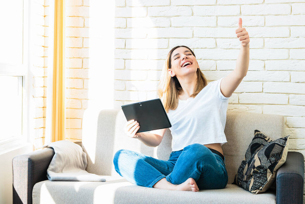 lucky beautiful woman on sofa at home holding a tablet and big thumb up. key to the success - Fotoğraf, Görsel