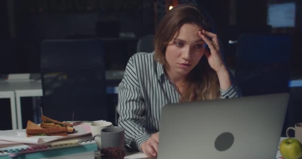 Tired woman working on laptop alone in dark office late at night. Stressed girl proping head, touching and wrying face while finishing urgent work in empty office . Concept of overwork. - Metraje, vídeo