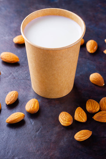Almond milk with almond on dark background. Homemade organic almond milk in a paper cup for healthy breakfast. Vegan milk from almonds nuts in kraft paper cup. Alternative milk. Zero waste - 写真・画像