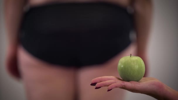 Close-up of female Caucasian hand holding apple as obese woman with cellulite standing at the background. Healthy food, dieting, overweight problem. - Πλάνα, βίντεο