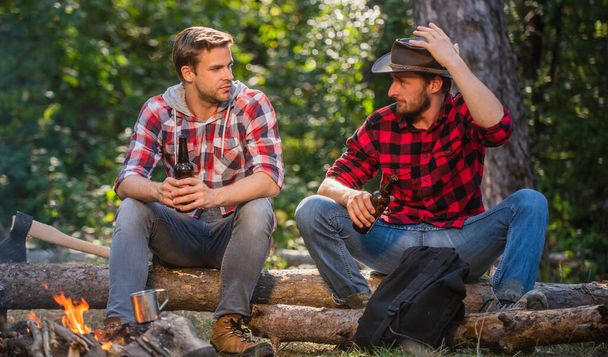 nice speak. man drink beer at bonfire. ranger at outdoor activities. spend picnic weekend in nature. Adventure concept. hike and people. two men relax at fire. hiking and camping. male friendship - Foto, Imagem