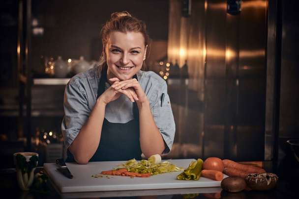 Lächelnde Köchin steht in einer Küche neben Schneidebrett, Schürze und Hemd, posiert für die Kamera, Reality-Show-Look - Foto, Bild