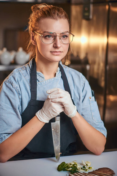 Pensando e pacífica chef feminino de pé na cozinha do restaurante, segurando uma faca contra a tábua de corte olhando pacientemente para a câmera
. - Foto, Imagem