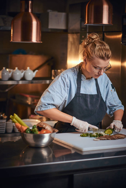 Europeo chef donna in piedi in una cucina accanto al tagliere, indossando grembiule e camicia, in posa per la fotocamera, reality show look
 - Foto, immagini