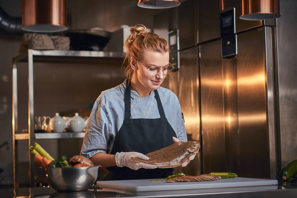 Interested female chef standing in a kitchen next to cutting board, holding a fish, wearing apron and shirt, posing for the camera, reality show look - Zdjęcie, obraz