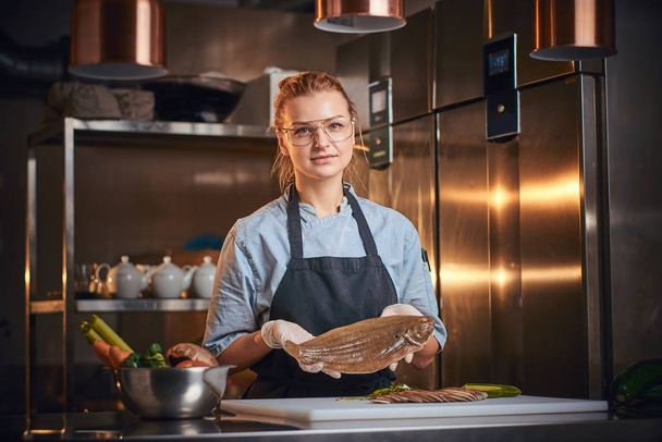 Interested female chef standing in a kitchen next to cutting board, holding a fish, wearing apron and shirt, posing for the camera, reality show look - Fotoğraf, Görsel