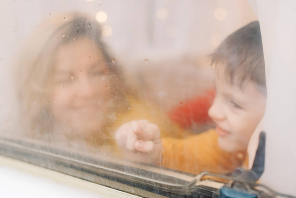 Όμορφη γυναίκα και ο χαριτωμένος γιος της κοιτάζουν έξω από το παράθυρο στο τρέιλερ. Ημέρα της Μητέρας ευχετήρια κάρτα - Φωτογραφία, εικόνα