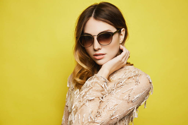Isolated portrait of a beautiful blonde young woman in sunglasses and cocktail dress over yellow background. Model girl in a trendy outfit posing at the yellow background - Photo, image
