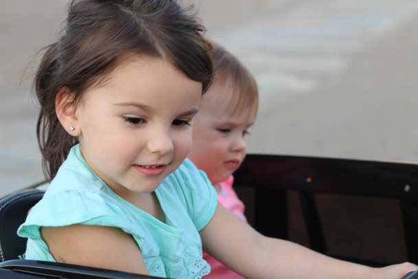 İki harika kız, asfaltta büyük bir oyuncak arabaya biniyor. Çocuklar için yaz eğlencesi olarak açık havada araba kullanmak - Fotoğraf, Görsel